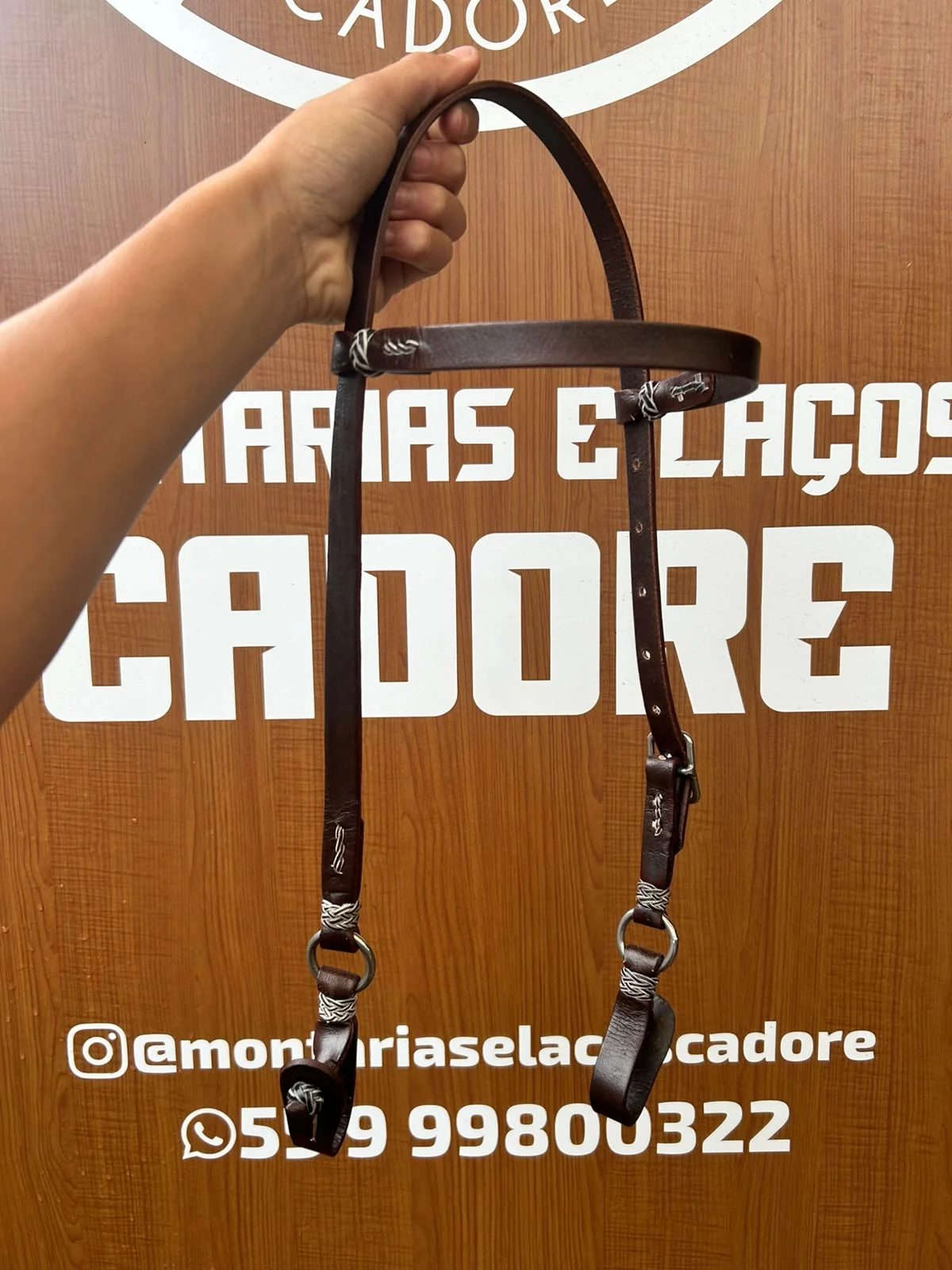 Cabeçada de couro marrom escuro segurada pela mão em frente ao painel da loja