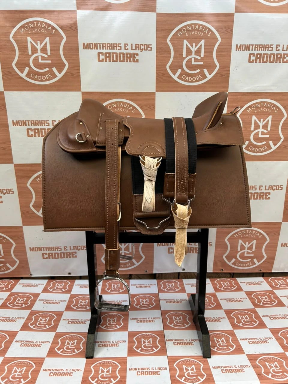 Kit de sela marrom montado no cavalete, com sincha preta, amarrilhos de couro cru e estribo