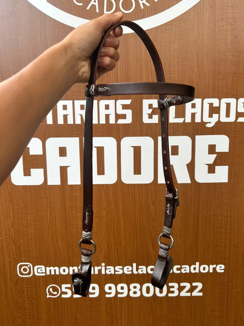 Cabeçada de couro marrom escuro segurada pela mão em frente ao painel da loja