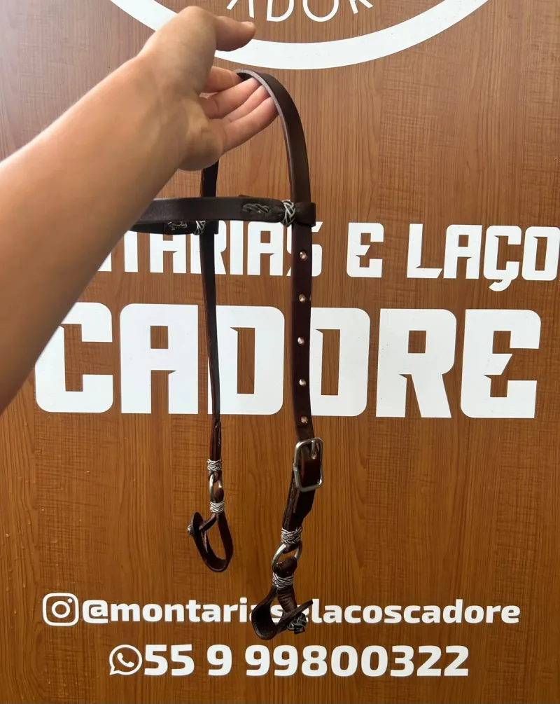 Cabeçada de couro marrom escuro com argola e ferragem, segurada pela mão em frente ao painel da loja